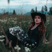 A woman with auburn hair wearing a black dress and cowboy hat sat in a field